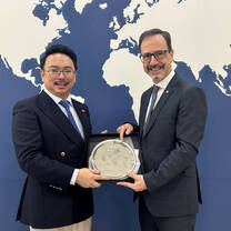 	 Two men in suits stand in front of a world map backdrop, smiling and holding a silver award plate together.