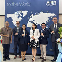 Six people stand in front of a world map backdrop at AIHM College, holding glasses and dressed in business attire. Two large floral arrangements are on either side of the group.