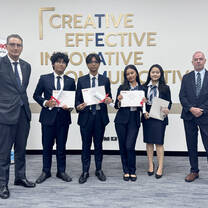 Five students in formal attire stand holding certificates and trophies, flanked by two men in suits. Behind them, a wall reads Creative Effective Innovative Solutions. Banners with educational information are to the left.