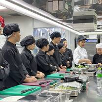 A group of chefs in black uniforms stand around a kitchen counter, attentively watching two instructors in white coats demonstrate a cooking technique with ingredients and utensils laid out in front of them.