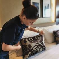 A woman in a dark uniform arranges decorative pillows on a neatly made bed in a well-lit, modern hotel room.