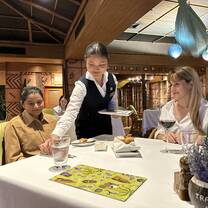 A waitress serves food to two women sitting at a restaurant table set with drinks and a colorful placemat. The setting is warmly lit with wooden decor and hanging blue lamps in the background.