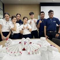 A group of six young adults stands behind a table decorated with towel art shaped like swans and flowers. They are smiling and wearing matching uniforms in white and navy blue, posing in a modern room.