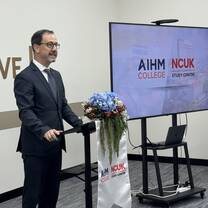 A man in a suit stands at a podium decorated with a blue floral arrangement, speaking in front of a screen displaying AIHM College and NCUK Study Centre logos in a classroom setting.