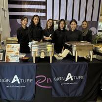 Six women in black uniforms stand behind a catering table with food warmers and promotional banners labeled "SIGNATURE" at an indoor event.