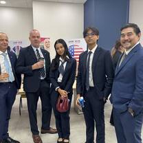 Five people in business attire stand together and smile at an indoor event. A table with refreshments and posters, one featuring the US flag, are in the background. Two people hold drinks and one carries a bag.