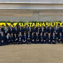 A group of students in formal attire pose together in front of a large "SUSTAINABILITY" sign with greenery in the background.