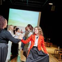 Students in costume smile at one another as they perform a choreographed dance in the centre of a film studio. behind them is a projection screen showing blue, yellow and green colours.