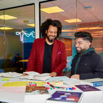 A professor and a students sit smiling at a table covered with various business books and study materials.