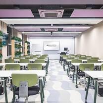 A spacious classroom with purple accent walls, a large wall-mounted screen, rows of tables, green and white chairs, geometric carpet and faux greenery.