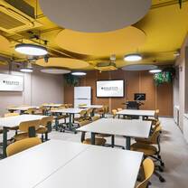 A spacious classroom with yellow accent walls, large wall-mounted screens, group tables, yellow accent chairs and faux greenery.