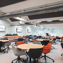 A spacious classroom with light blue walls, large wall-mounted screens, group tables, chairs of various colours and a patterned carpet.