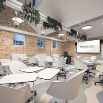 A spacious classroom with exposed brick walls, a large wall-mounted screen, white group tables, white chairs and a patterned carpet.