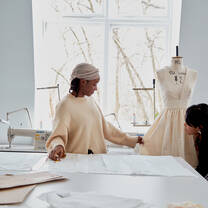 A student and a professor are working on a mannequin in a bright, white-walled fashion design studio. There are sewing machines on the background and a large table covered in sheets of paper with sketches.