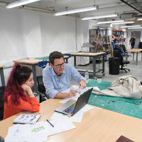 A student and a professor sit at a table with a laptop in front of them. They are in a basement room which has several worktables, green cutting maps and model structures in it.