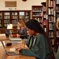 A student sitting in a library at a long table with her laptop in front of her and several shelves of books in the background.