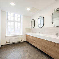 A bright white-walled bathroom with large white windows, grey wood flooring, oak and white basis and black oval mirrors
