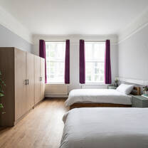 A bright bedroom containing two neatly made beds set in front of two large white windows and opposite two oak wardrobes.