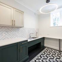 A bright kitchen with white walls, black and white geometric tiles floors and deep green kitchen units with white countertops.