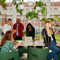 A group of students standing around a long table in front of a large white-pane window. The students are looking through text books, working on laptops, drinking coffee and chatting and laughing with one another. 