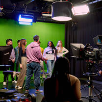 A group of students stand in front of a green screen in a TV studio smiling and discussing with one another. Towards the front, there is a student holding a clapperboard. Opposite them is a student standing behind a large teleprompter and camera.