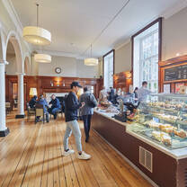 A spacious coffee shop with white painted and wood walls, wood flooring and a display case on the right hand side showcasing various cakes and pastries.