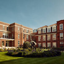 A brick three-floor building with several white windows overlooks central square with a green lawn and sculpture in the centre.