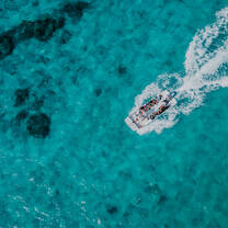 Aerial view of a small boat speeding across bright turquoise water, leaving a white wake behind it as people ride on board.