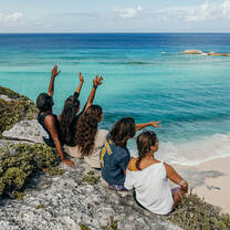 Five people sit on a rocky cliff overlooking turquoise ocean water, some with arms raised or pointing toward the horizon, enjoying the coastal view on a sunny day.