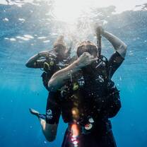 Two scuba divers underwater facing the camera, making hand signals as sunlight filters through the surface above them.