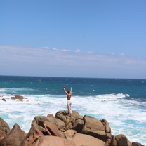 A girl standing on a rock by the sea with her hands in the air looking out towards the water. Taken from afar.