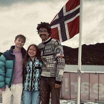 three college students stand smiling with the Norwegian flag waving in the wind