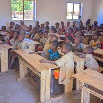 A schoolroom of pupils looks on as a woman reads from a packet.