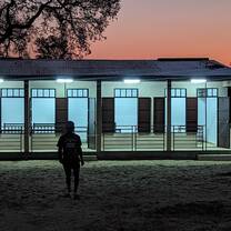 The sun sets behind the porch of a white stucco building, a visitor silhouetted in front of it