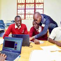 A teacher supports three upper-primary school students using laptops during a technology class in a classroom setting.