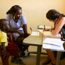 A volunteer meets with a parent and young child at a table, reviewing notes during a community based health consultation.