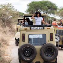 A young girl wearing a sun hat stands at the back of an open-top safari vehicle in Tanzania, looking through binoculars.