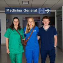 Three medical interns wearing blue scrubs stand together smiling beneath a 'Medicina General' sign in a Peruvian hospital corridor.