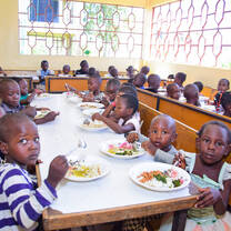 Young children seated at long tables eating a prepared lunch inside the Maisha Academy dining hall as part of the daily school meal program.