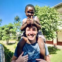 Volunteer carrying a young child on their shoulders outdoors at the Maisha campus, both smiling during daily activities.