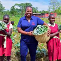 A staff member and two primary school students hold baskets of leafy vegetables harvested from the school farm.