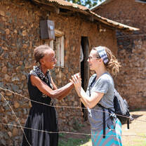A volunteer and an elderly community member greet each other outside a rural home during a home visit.