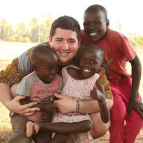 Volunteer kneeling outdoors with three young children, smiling and embracing them during a community program at Maisha.