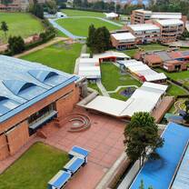 The image shows an aerial view of an educational institution with a spacious and well-organized campus, featuring brick buildings with blue and white roofs, well-maintained green areas, recreational spaces, and sports facilities such as an athletic track. Walkways and courtyards connect the different areas, and the surroundings combine nature with urban buildings, conveying a sense of order, openness, and institutional well-being.