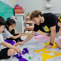 Male TEFL teacher kneels on the floor to help students with their task.