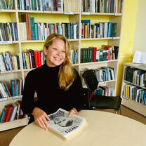 Smiling female TEFL teacher sat at table in front of a bookshelf holding some papers.
