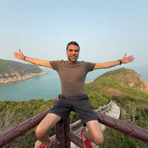 Man sitting on a viewpoint railing with arms outstretched, overlooking a coastal landscape.
