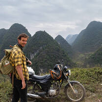 A man with a backpack smiles next to a motorcycle, with a backdrop of steep, lush green mountains under a cloudy sky in Vietnam..
