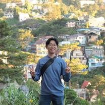 A man in a blue shirt makes peace signs against a backdrop of a colorful hillside town and green trees.
