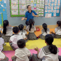 TEFL teacher leading a classroom of young students seated on the floor, using hand gestures during a lesson.